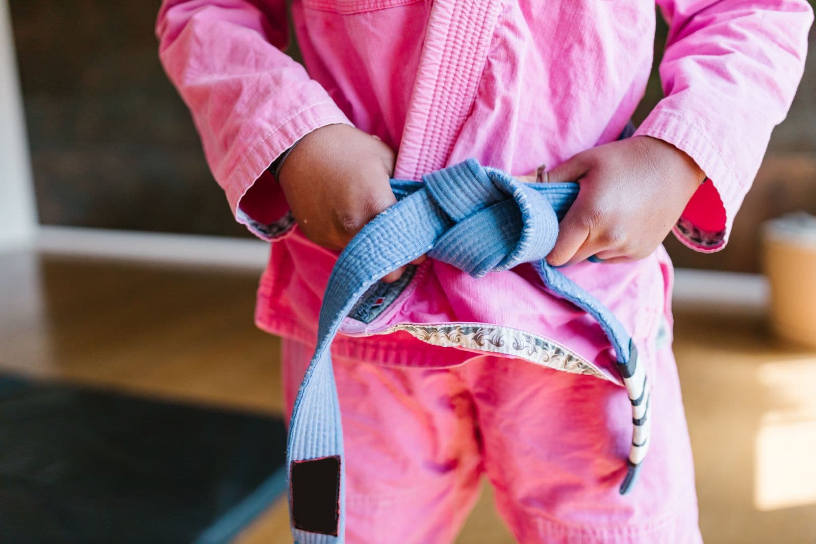 A child in a pink martial arts uniform tying a blue belt, seen from the chest down