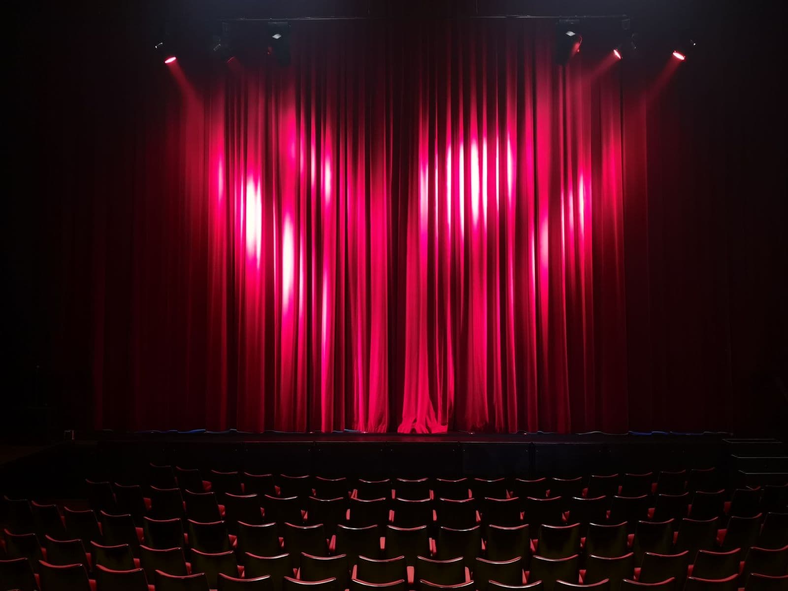 An empty theatre stage with red curtains under spotlights, seen from the seats