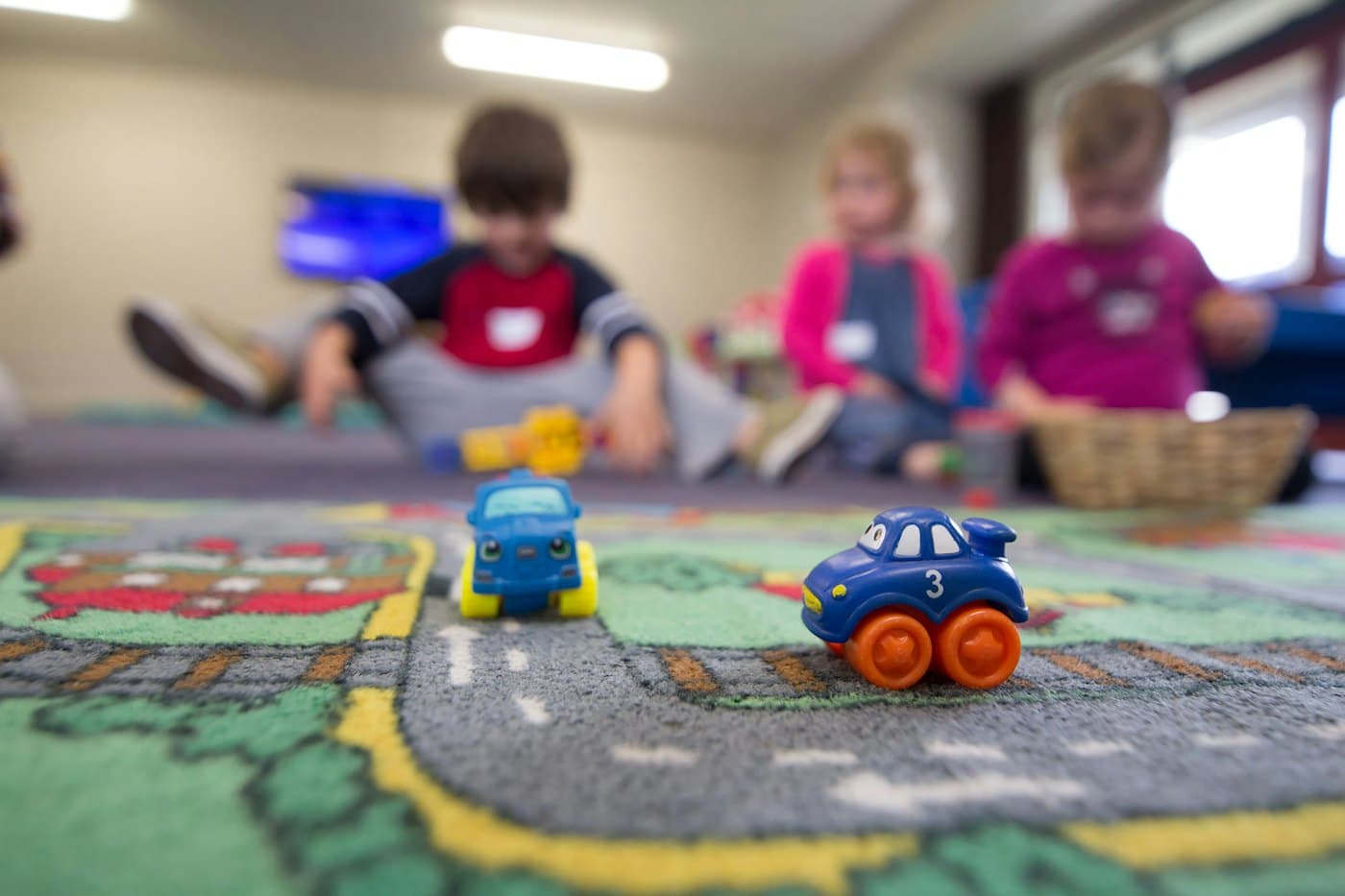 Toy cars on a play mat while children play in the background