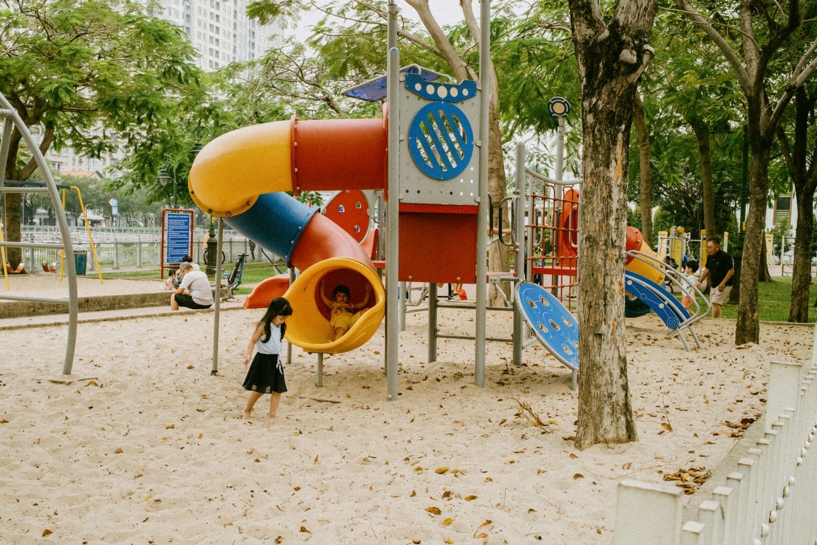 Colourful slides and climbing structures on a sandy park playground, with children playing in the distance