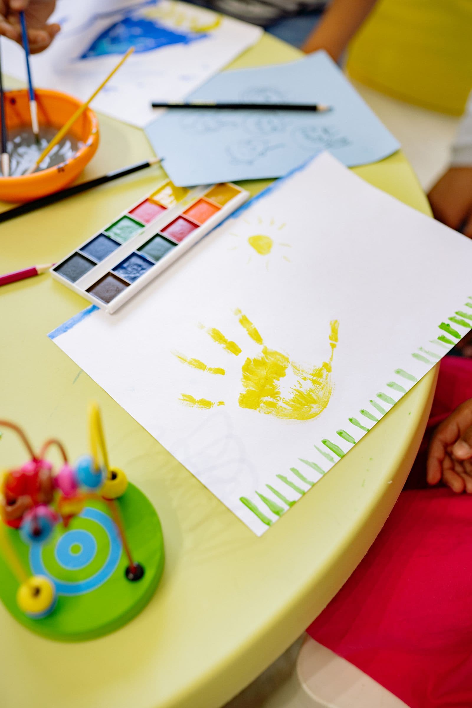 A yellow handprint painting beside watercolours and a bead toy on a preschool table