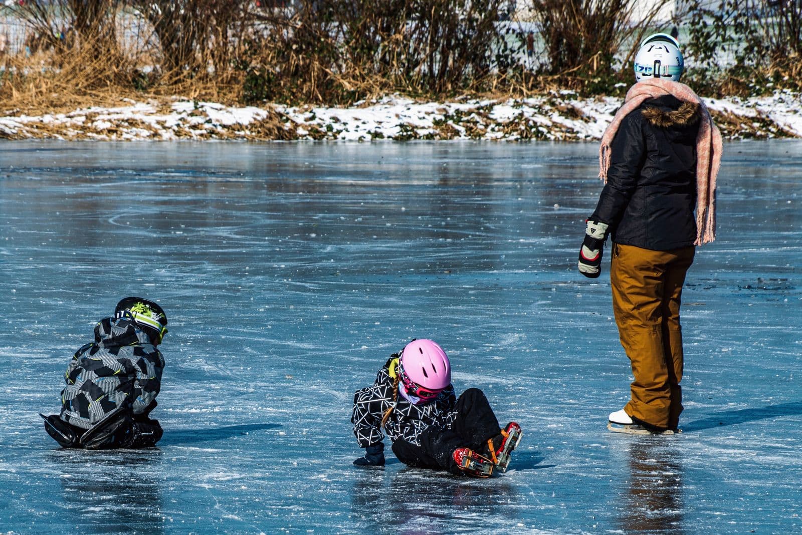 Two young children in helmets and skates on the ice, with an adult skating beside them, all seen from behind