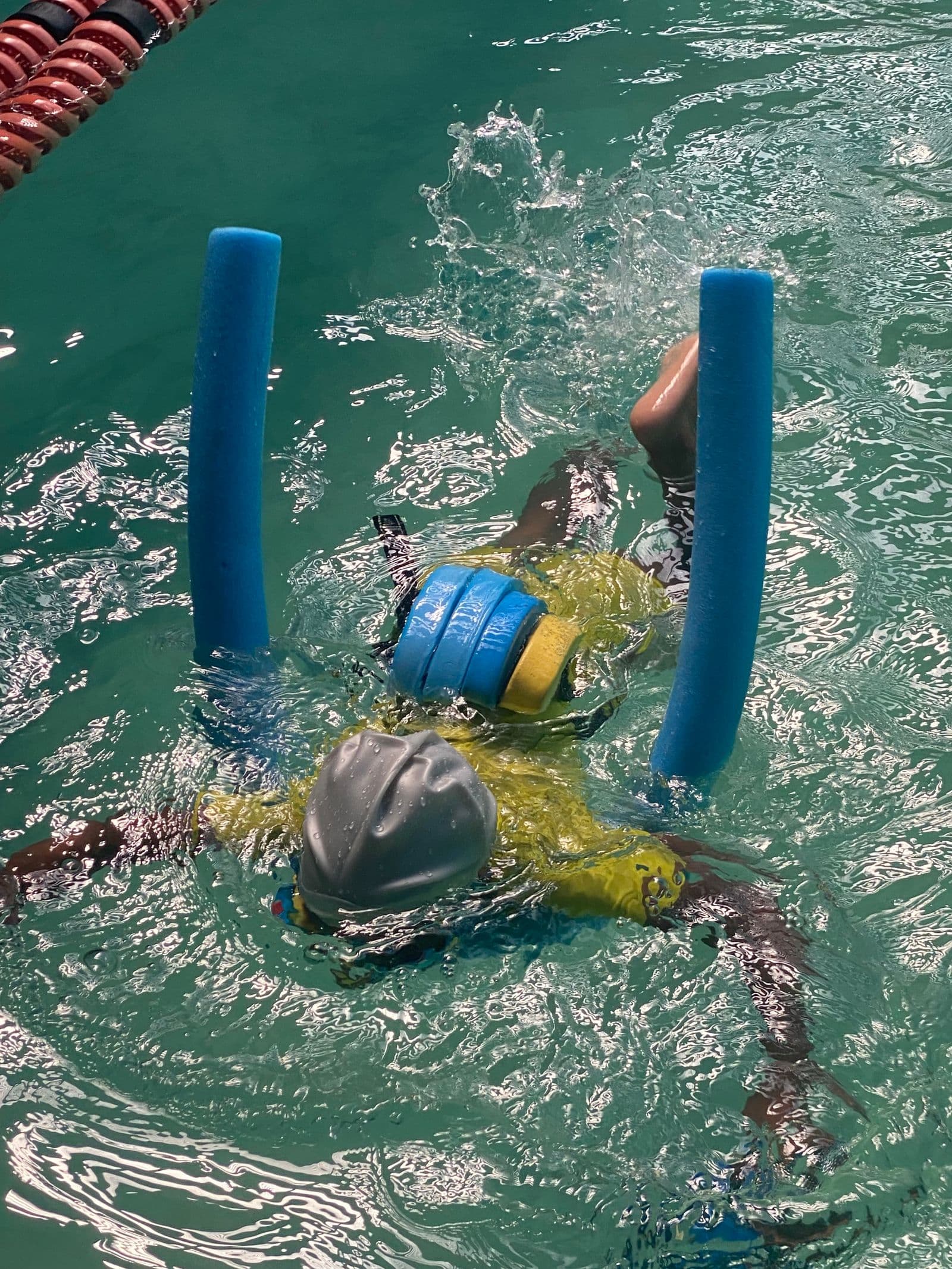 A child in a swim cap and float belt, face down in the water, learning to swim with pool noodles